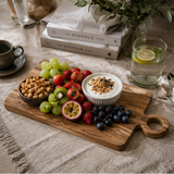 Fruit platter with yogurt and nuts on a wooden board, set on a table with books and glasses.