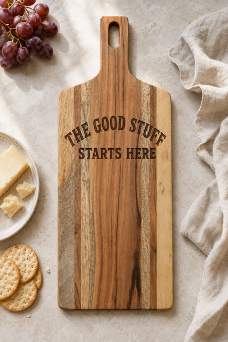 A rectangular wooden serving board with a handle, featuring the engraved text 'THE GOOD STUFF STARTS HERE' in dark brown lettering. The board is styled in a flat-lay arrangement on a light beige textured surface, surrounded by food props including grapes, crackers, cheese, and a beige cloth.