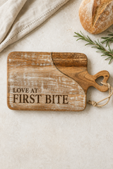 A rustic wooden cutting board with the phrase 'LOVE AT FIRST BITE' engraved on it, styled on a light beige surface with a loaf of bread, a sprig of rosemary, and a folded cloth.