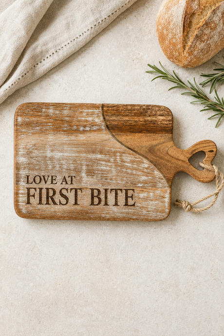 A rustic wooden cutting board with the phrase 'LOVE AT FIRST BITE' engraved on it, styled on a light beige surface with a loaf of bread, a sprig of rosemary, and a folded cloth.