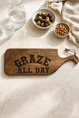 A rustic wooden serving board with the words 'GRAZE ALL DAY' engraved in dark lettering, styled on a light marble surface with olives, almonds, glasses, and a cloth napkin.
