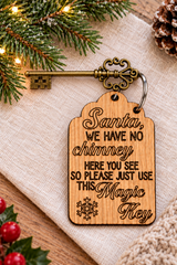 A wooden Christmas key tag engraved with the phrase 'Santa, WE HAVE NO chimney HERE YOU SEE SO PLEASE JUST USE THIS Magic Key' and a snowflake, shown with a decorative antique-style metal key on a burlap fabric with holiday greenery and lights.