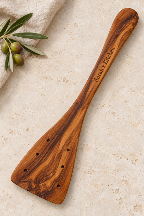 Olive wood spatula with six round holes and “Sarah’s Kitchen” engraved on the handle, beside linen and olives.