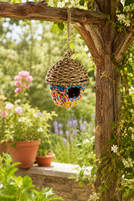 Colorful woven birdhouse hanging from a tree branch in a garden setting.