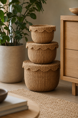 Three nested round woven baskets with scalloped edges in a natural tan jute or seagrass material, styled on a rug next to a wooden cabinet and a potted plant.