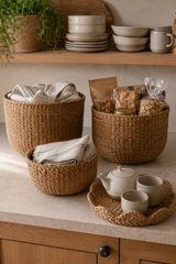 Three round woven wicker baskets in different sizes arranged on a light stone countertop in a warm kitchen setting, with folded linens, packaged snacks, and a woven tray holding a teapot and cups.