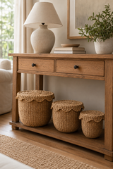 A rustic wooden console table styled in a bright living room, holding a cream ceramic table lamp, stacked books, a potted green plant, and three woven natural fiber storage baskets on a lower shelf.
