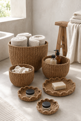 A coordinated set of woven natural-fiber baskets and matching tray-like holders arranged in a bright bathroom setting, with rolled towels, soap dispensers, and a bar of soap inside.