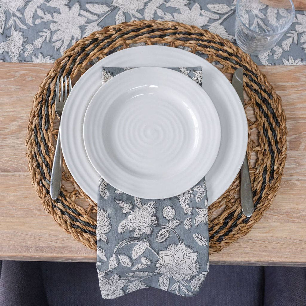 Set table with white plates on a round handwoven seagrass placemat with a natural two-tone spiral weave and scalloped edges, silverware, and a floral-patterned napkin on a wooden table.