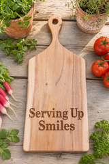 A rectangular wooden serving board with a handle, engraved with the words 'Serving Up Smiles', displayed on a rustic wooden table surrounded by fresh radishes, tomatoes, and potted herbs.