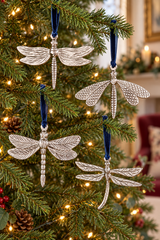 Four silver dragonfly ornaments with textured wings and blue ribbons hang on a decorated Christmas tree with warm lights.