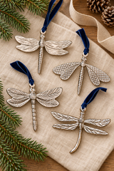 Four dragonfly-shaped hanging ornaments in textured silver-tone metal with dark blue velvet ribbons, arranged on a beige fabric surface with pine branches and a ribbon in the background.