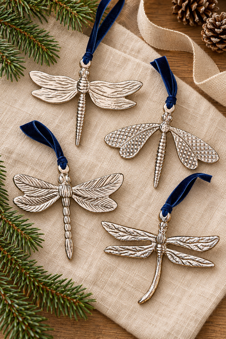 Four dragonfly-shaped hanging ornaments in textured silver-tone metal with dark blue velvet ribbons, arranged on a beige fabric surface with pine branches and a ribbon in the background.