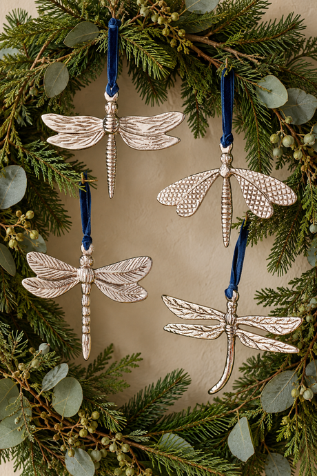 Four silver dragonfly ornaments with blue ribbons, hanging from a green eucalyptus wreath against a neutral beige background.
