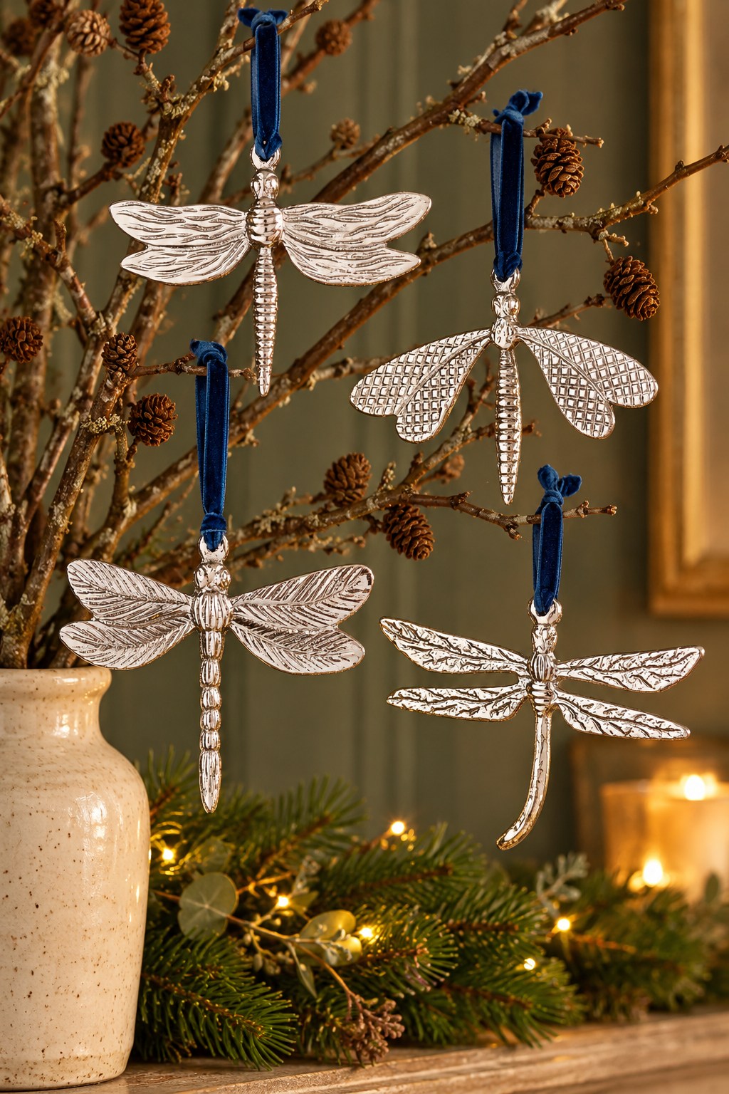 Four silver dragonfly ornaments with blue hanging ribbons displayed on decorative branches in a vase, with a holiday garland and warm lights in the background.