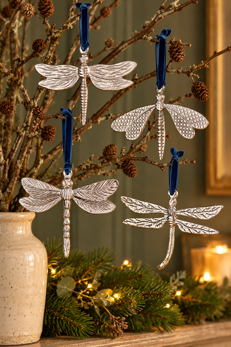 Four silver dragonfly ornaments with blue hanging ribbons displayed on decorative branches in a vase, with a holiday garland and warm lights in the background.