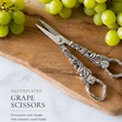 Silver grape scissors with decorative design on a wooden board with grapes and cheese.