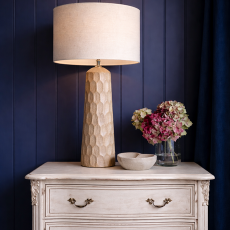 Vintage-style chest of drawers with a lamp and flowers against a dark blue wall.