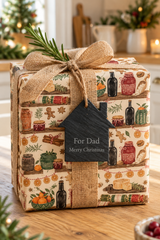 A square gift box wrapped in a warm-toned Christmas kitchen-themed patterned paper, tied with a burlap ribbon and topped with a pine sprig. A small black house-shaped tag reads 'For Dad Merry Christmas.