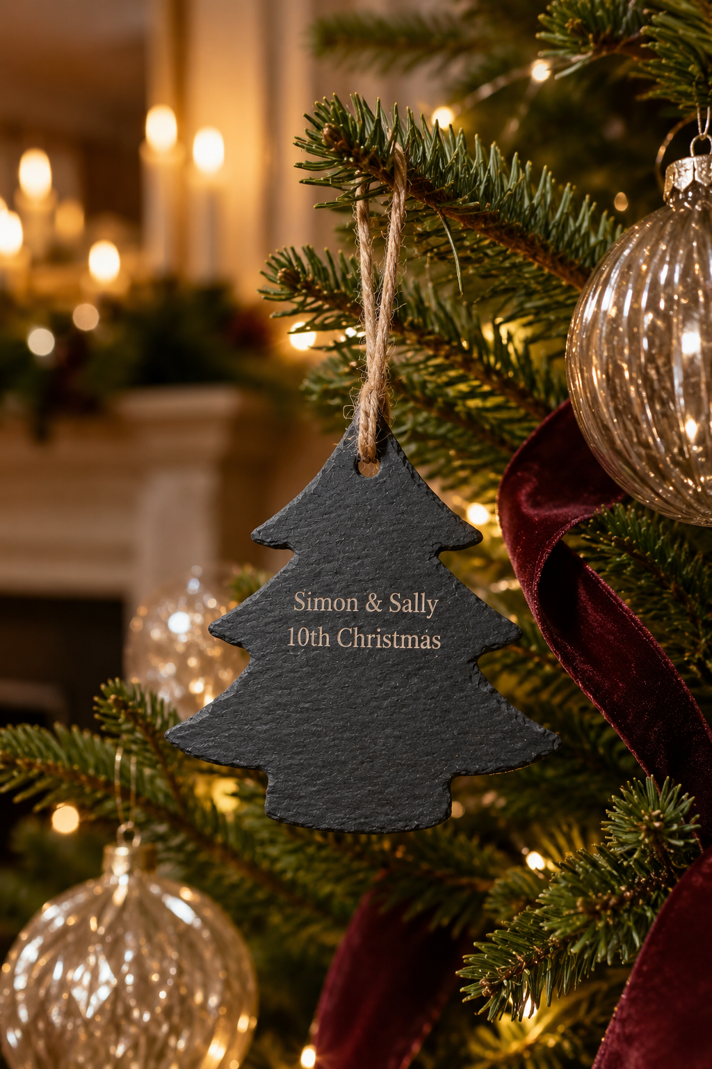 A personalized Christmas tree ornament shaped like a dark slate Christmas tree, hanging on a decorated tree branch with warm lights and glass ornaments in the background.