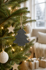 A dark gray slate Christmas tree ornament hanging on a decorated green Christmas tree in a cozy living room setting with warm lights and wrapped gifts.