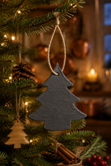 A dark gray slate Christmas tree ornament hanging from a green pine tree branch with warm string lights in the background.