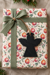 A wrapped Christmas gift box with a festive pattern of red ornaments, green holly leaves, and gold stars, tied with an olive green ribbon and bow. A black slate Gift Tag angel silhouette hangs from the ribbon on a beige linen background with pine branches at the top and bottom corners.