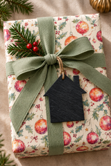 A wrapped Christmas gift box with festive wrapping paper featuring red ornaments and green holly motifs, tied with a wide sage green ribbon bow and decorated with a pine sprig and small red berries. A black house-shaped tag hangs from the ribbon on a beige fabric background.