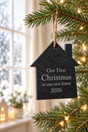 A house-shaped black slate Christmas ornament hanging from a pine tree branch with warm string lights. The ornament is engraved with the text 'Our First Christmas in our new home 2026' in white lettering.