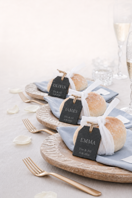 Personalised Slate Tags wedding favour - name plate on Three rustic bread rolls on blue napkins with white ribbon. Laser engraved place cards, arranged on round wooden plates with gold forks on a light tablecloth.