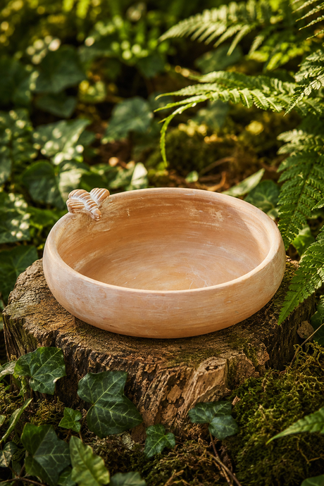 A round, handcrafted terracotta clay bowl with a small bee detail on the rim, used for bird bath, bird feeder displayed on a tree stump in a lush green forest setting.