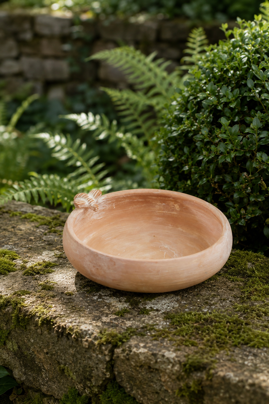 A shallow round terracotta clay bowl with a small bee detail on the rim, used for bird bath, feeder displayed on a mossy stone in a garden setting.