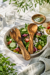 Small wooden salad servers made from solid olive wood. Salad with wooden utensils on a table with a white tablecloth and greenery.