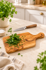 An olive wood cutting board with a handle, shown on a beige countertop in a kitchen setting. It is styled with fresh chopped parsley, a kitchen knife, a small bowl of coarse salt, and a striped cloth napkin.