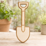 A small wooden garden shovel decoration standing upright on a light wooden surface, with a blurred background of potted plants.