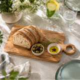 Loaf of bread on a wooden cutting board with olive oil and olives, set on a table with a glass of water and flowers.