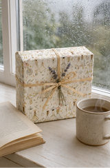 A gift box wrapped in cream-colored paper with a delicate Summer Fashion Sketch illustration pattern, tied with a natural twine ribbon and a sprig of dried lavender, sitting on a windowsill next to an open book and a ceramic mug.