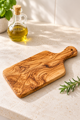 An olive wood cutting board with a natural grain pattern, styled on a light beige stone countertop next to a glass bottle of olive oil and a sprig of rosemary.
