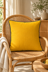 Plain mustard square cushion with blue piping against white pillows and a natural linen upholstered headboard.