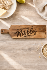 A wooden snack nibble board with the phrase 'BRING ON THE Nibbles' engraved on it, styled on a light wood tabletop with crackers, a pear, oats, and a napkin nearby.