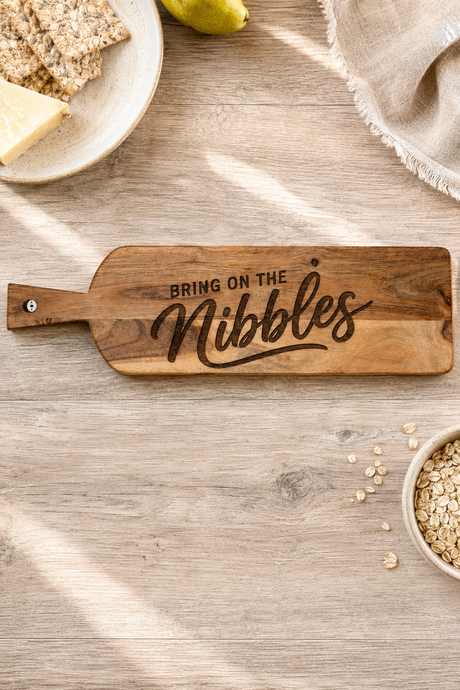 A wooden snack nibble board with the phrase 'BRING ON THE Nibbles' engraved on it, styled on a light wood tabletop with crackers, a pear, oats, and a napkin nearby.
