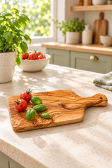 A rectangular olive wood cutting board with a handle, styled on a light beige marble kitchen countertop with fresh tomatoes and basil leaves. The background shows a softly blurred, bright kitchen with green cabinets and plants.