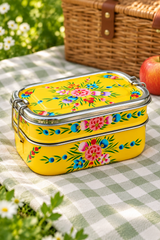 A bright yellow metal bento lunch box with colorful floral embroidery, resting on a green and white checkered picnic blanket with a wicker basket and apple in the background.