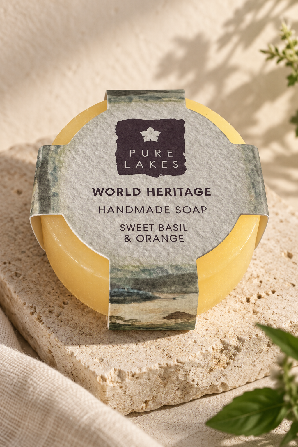 A round bar of Pure Lakes Sweet Basil & Orange handmade soap sits on a textured stone slab. The soap has a pale yellow wax body and a decorative paper label featuring a dark purple brand logo and a scenic coastal illustration.