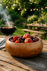 A Teak Root Fruit & Serving Bowl – 20cm. Round wooden bowl filled with fresh strawberries, peaches, and mixed berries on a rustic wooden table, with a blurred outdoor backyard scene in the background including a grill and string lights.