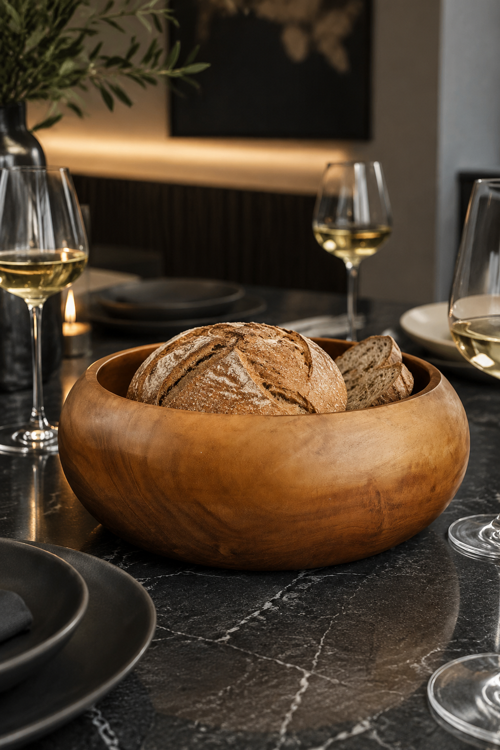 A Teak Root Fruit & Serving Bowl – 20cm. Round wooden bowl filled with rustic artisan bread sits on a dark marble table in a dining setting, surrounded by wine glasses and plates.