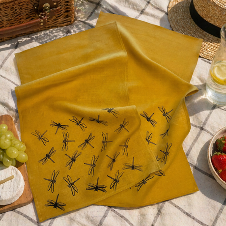 Yellow linen tea towel with black dragonfly patterns on a picnic setup with a basket, fruits, and a hat.