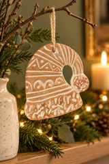 A hand-painted terracotta Christmas ornament in the shape of a Santa hat  decorated with white geometric patterns, hanging from a natural jute string on a decorative branch over a mantel.