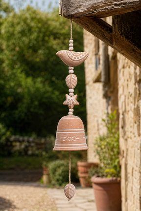 A rustic terracotta wind chime hanging from a wooden eaves in an outdoor garden setting. The wind chime features a bird, leaves, a star, and a bell with carved details, in a warm terracotta color.