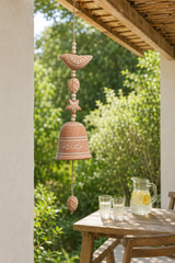 A terracotta-colored  wind chime hanging outdoors on a wooden porch, with a table, glasses, and a pitcher of lemonade in the background against a lush green garden.